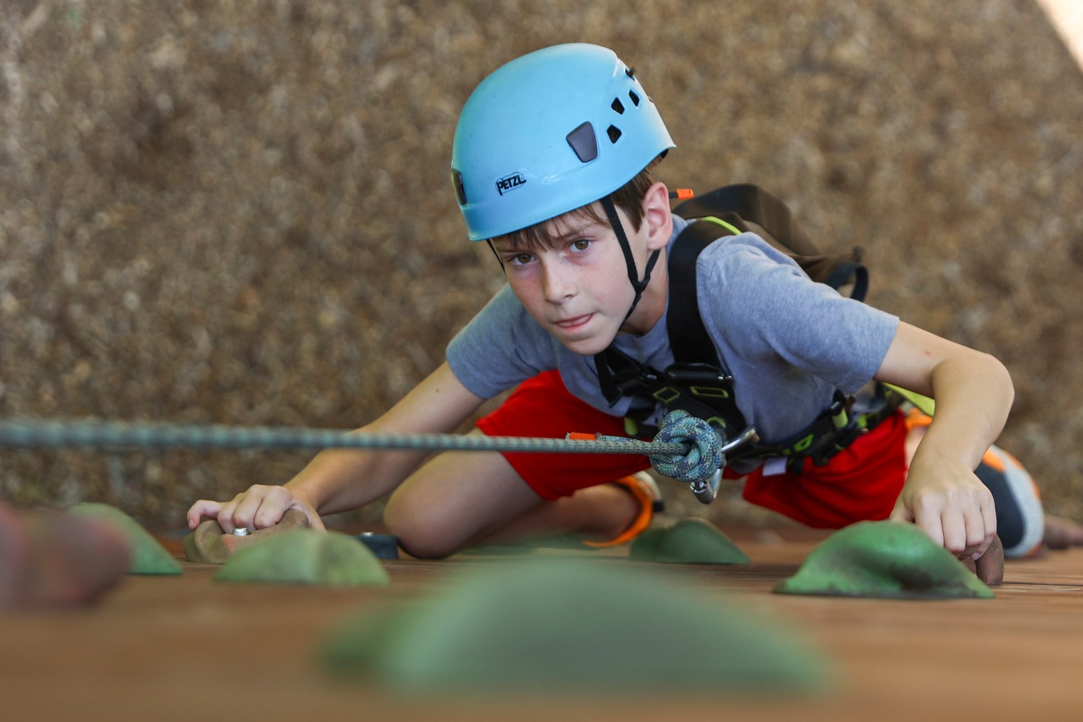 A boy wearing a safety harness and helmet climbs a rock wall, gripping the holds with determination.