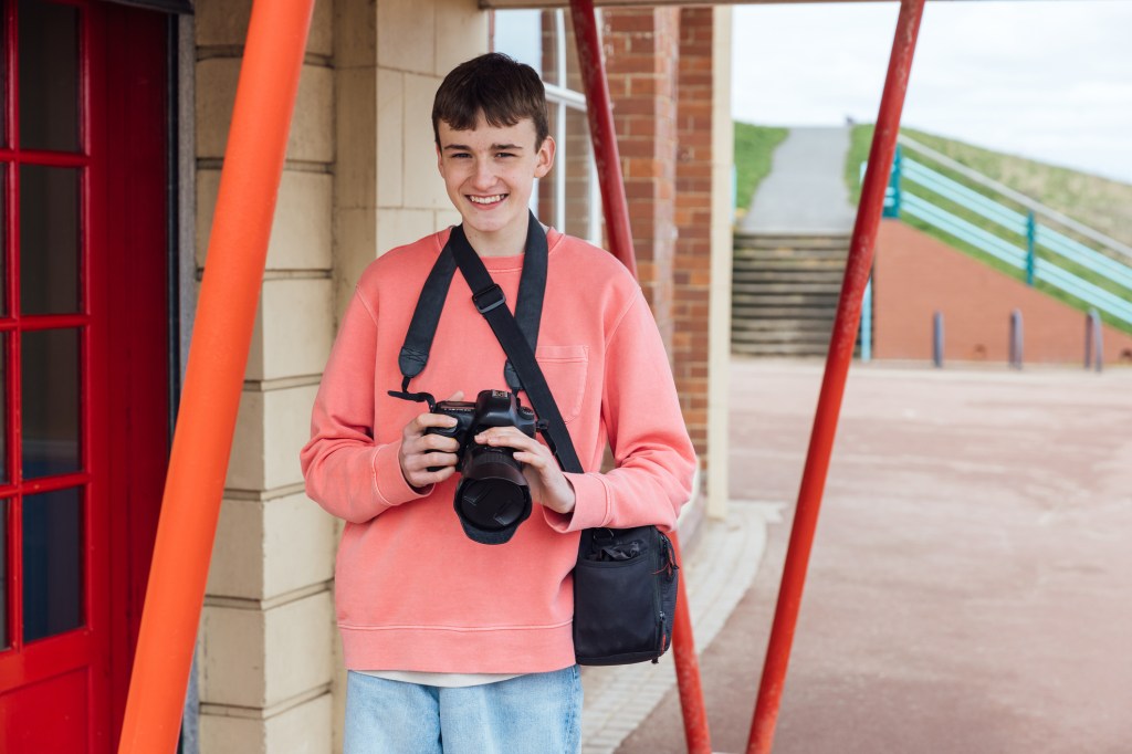 A smiling teen holding a DSLR camera stands outdoors near a building with bright red and orange structural beams, wearing a coral sweatshirt and a black camera bag across his shoulder.