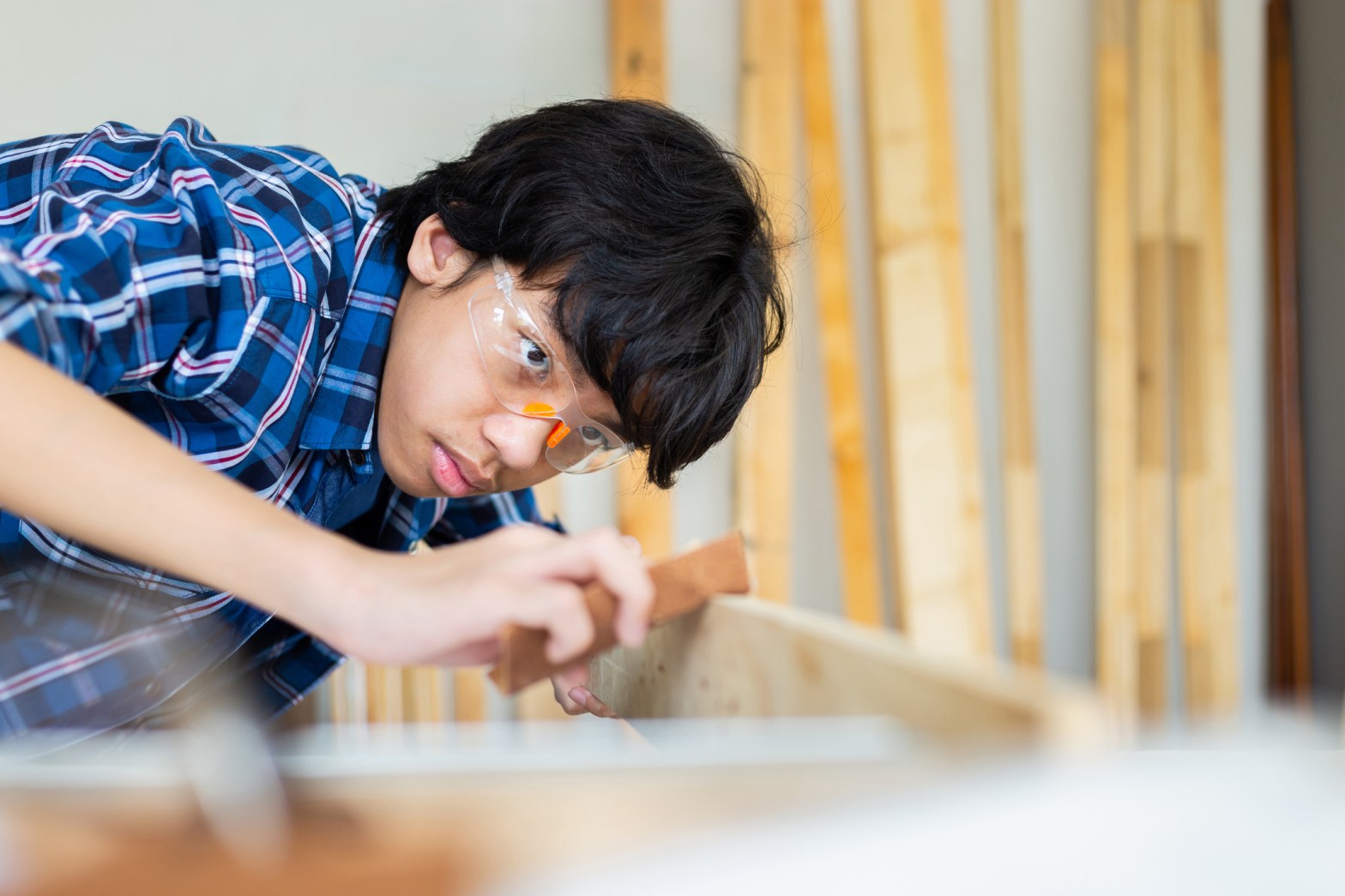 A young person wearing safety goggles sands a piece of wood, leaning in with focus while working on a woodworking project in a workshop filled with lumber.