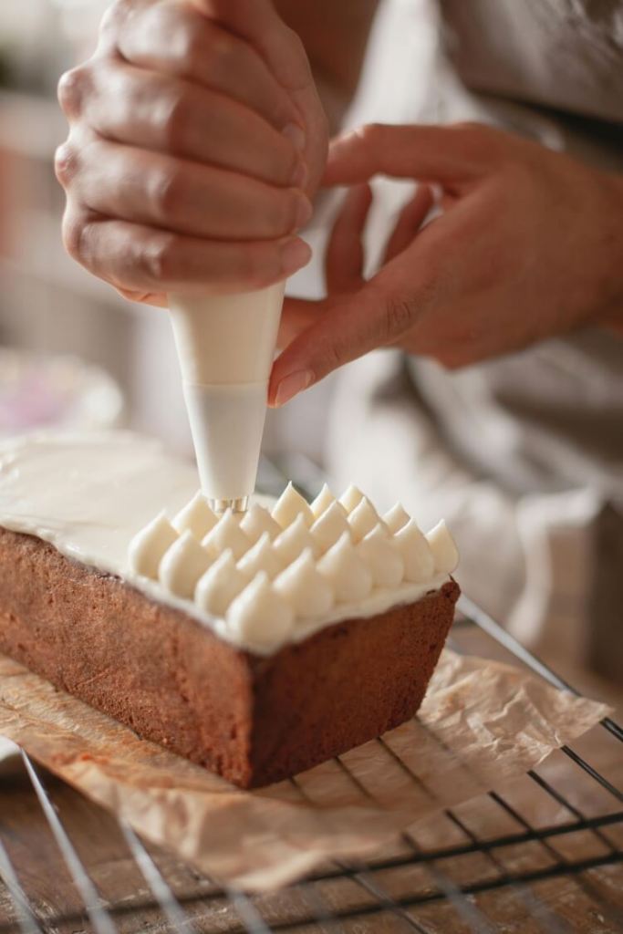 A person uses a piping bag to decorate a loaf cake with white frosting, creating peaks on top. The cake sits on parchment paper on a cooling rack.