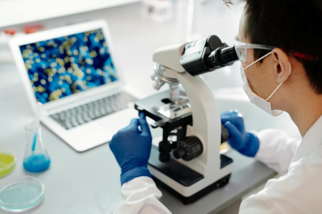 A scientist wearing a mask and gloves looks into a microscope at a lab bench, with a laptop displaying an image of colorful microorganisms or cells in the background.
