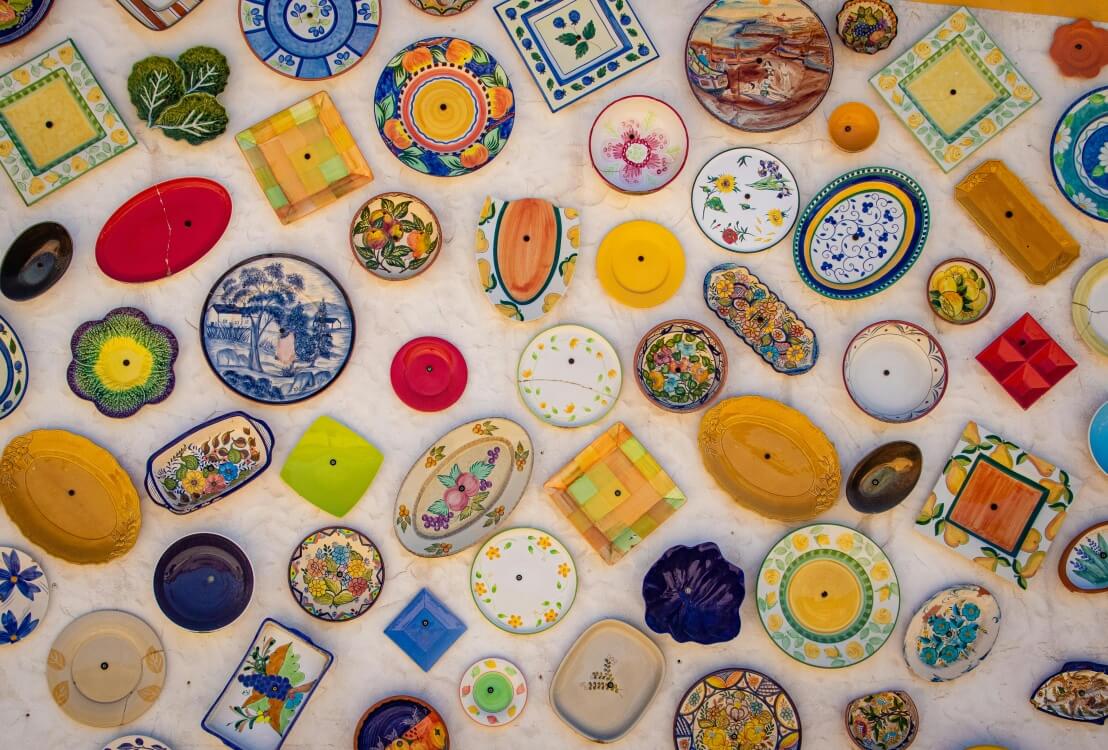 a wall covered in a vibrant collection of assorted, decorative ceramic plates. The plates vary widely in size, shape, and color, showcasing a diverse array of hand-painted designs and patterns
