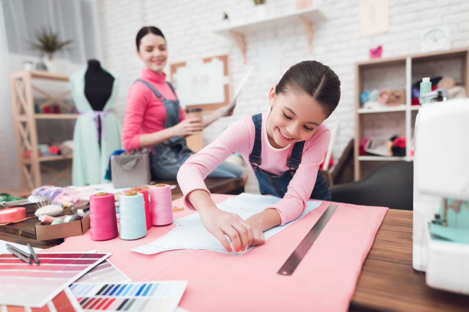 A smiling young girl pins a sewing pattern onto pink fabric at a craft table, surrounded by colorful thread spools, fabric swatches, and sewing tools, while an adult sits in the background holding a cup and watching her work.