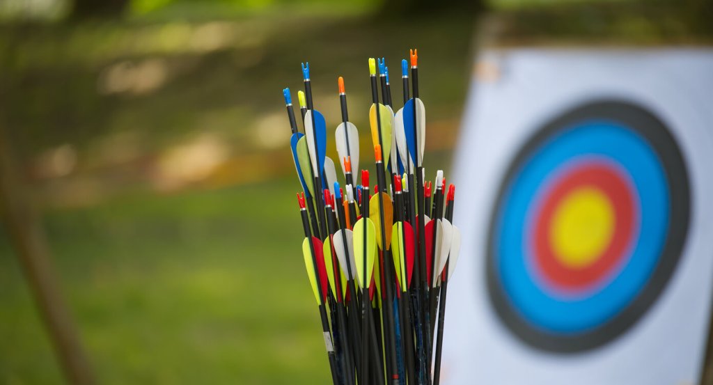 A bundle of colorful arrows standing upright in front of an archery target with concentric rings of blue, red, and yellow.