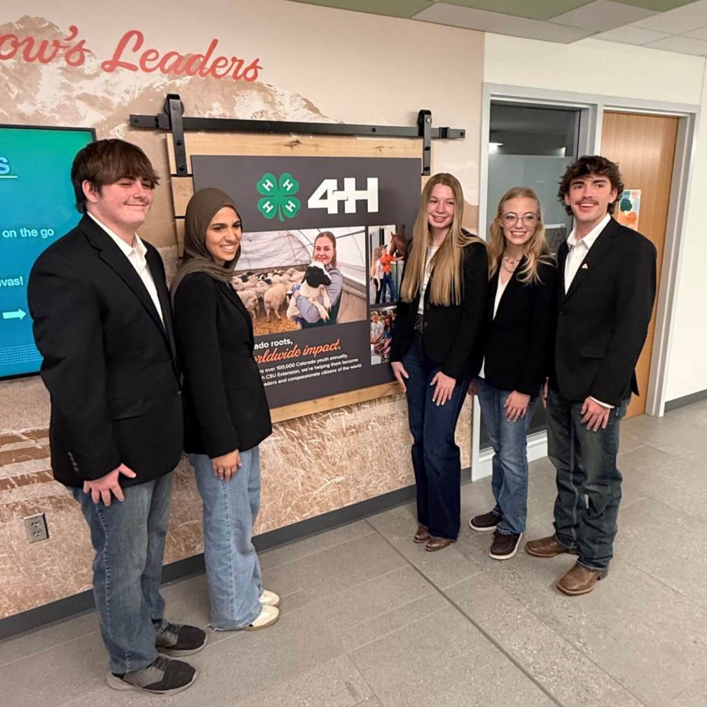 A group of Collegiate 4‑H members standing together in front of a large 4‑H display inside a building, all dressed in black blazers and jeans.
