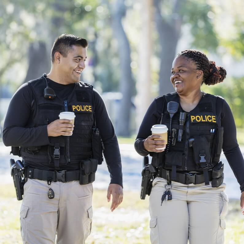 Two police officers wearing tactical vests walk side by side outdoors, each holding a takeaway coffee cup.